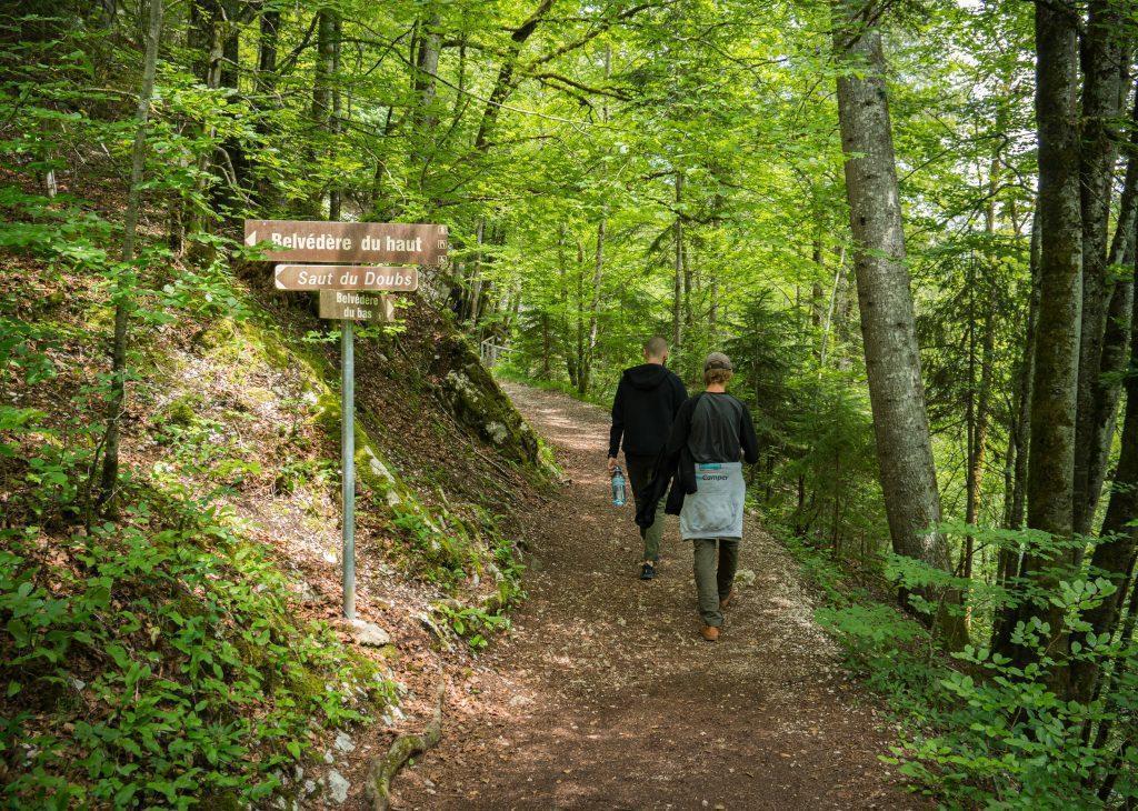 Zwei Männer gehen im Wald in Richtung Wasserfall-Doubs, davor Wegweiser - Wandern Jura